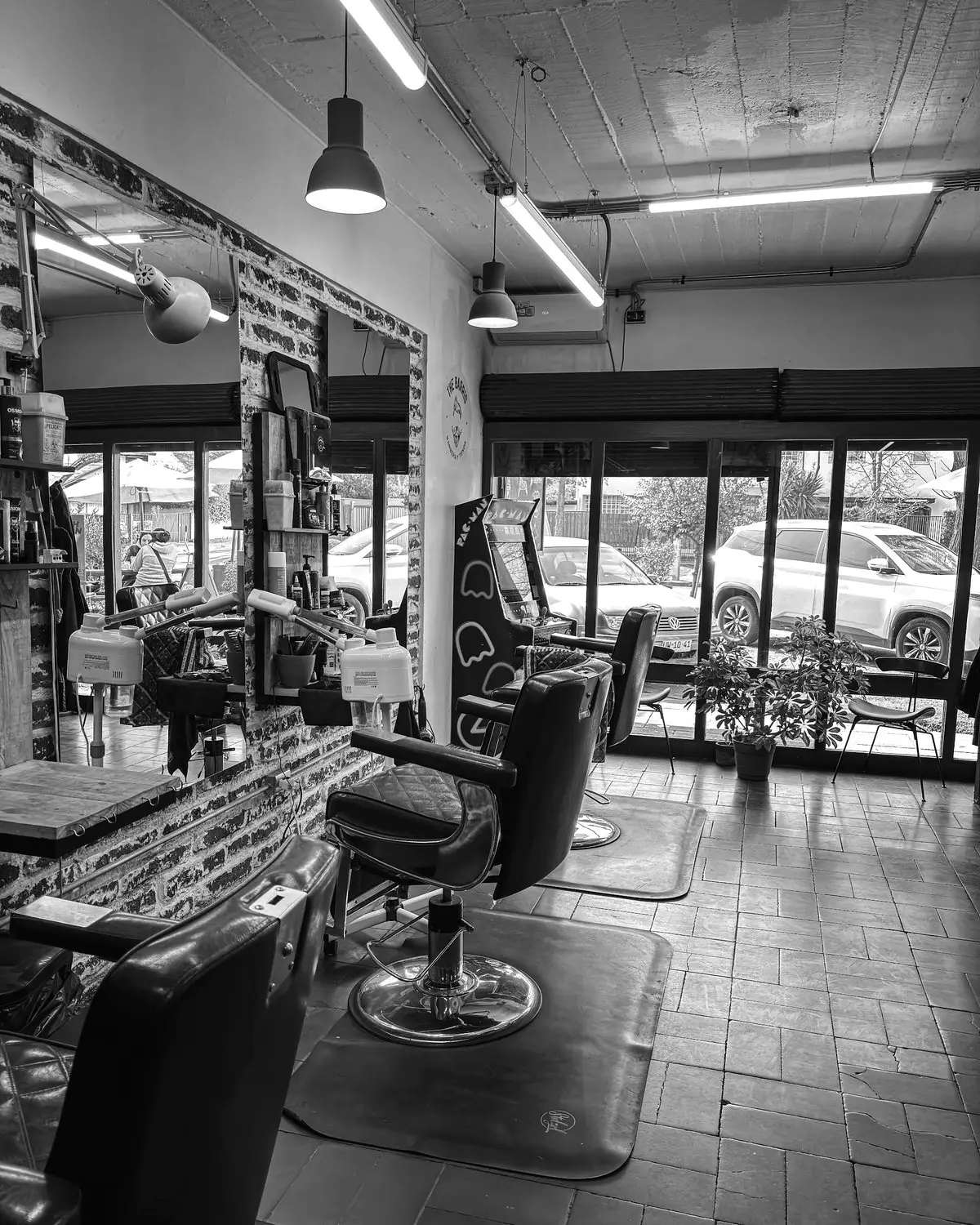 El salón de la barbería: sillones de barbero antiguos frente a un muro de ladrillo con espejos, techo de hormigón con luminarias, ventanal a la calle y una máquina arcade. En la pared del fondo, la chapa del logo.